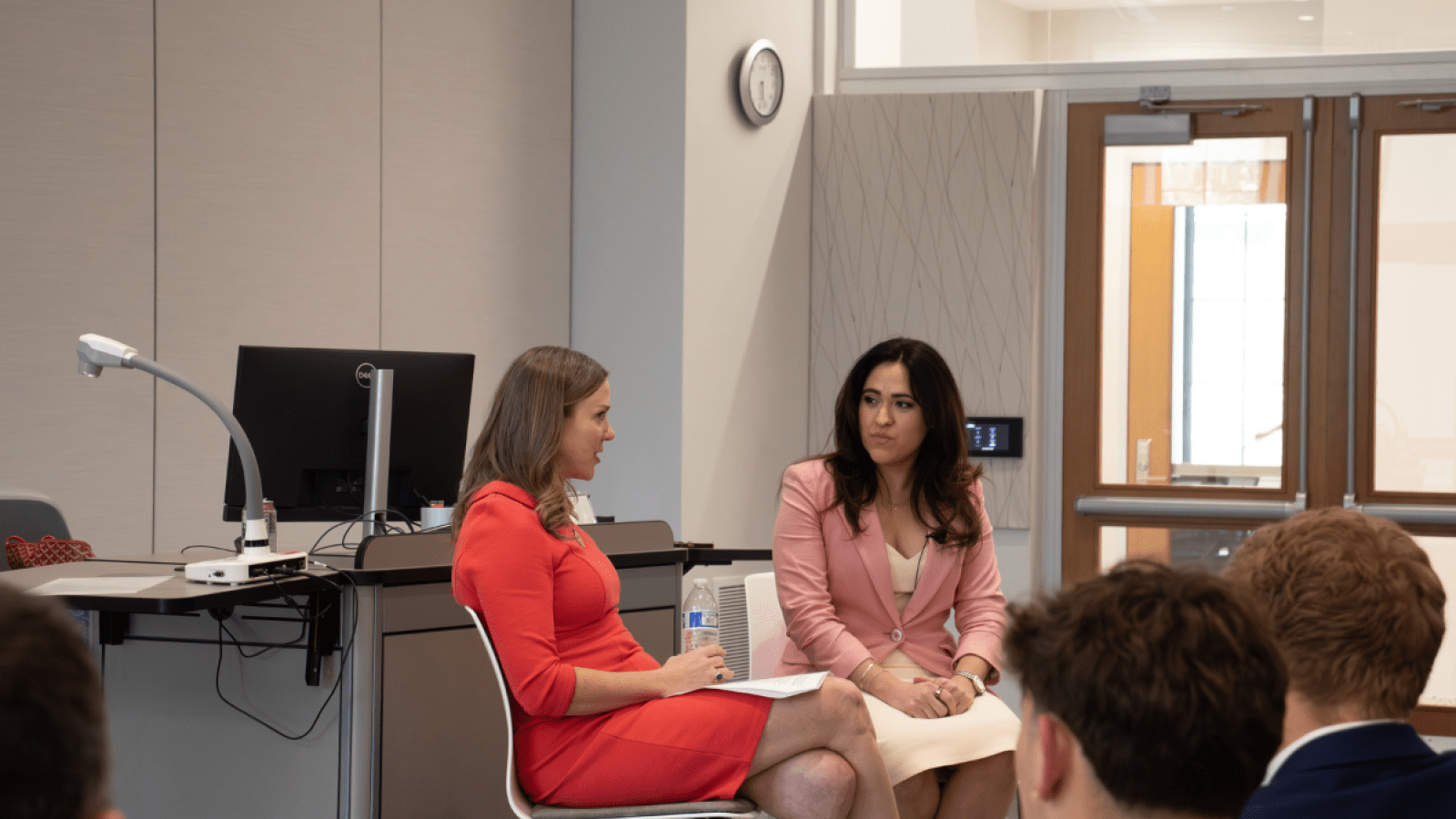 Two women are speaking to each other during a conversation in a classroom. One is wearing a red dress, another is wearing a pink blazer over a white dress.