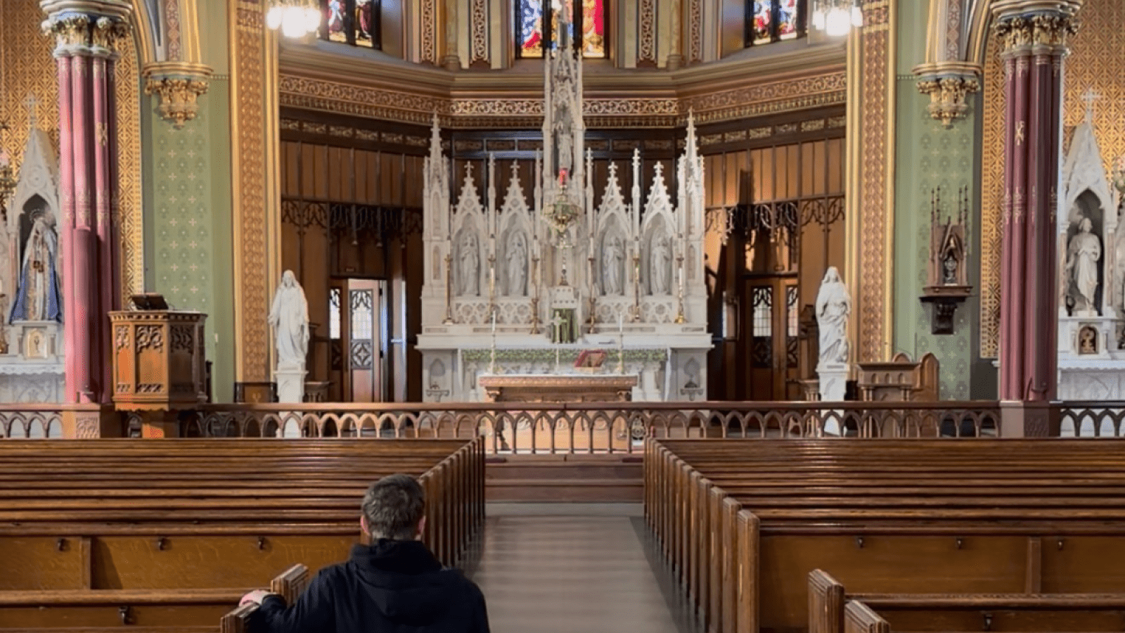 Grady Connolly genuflecting at the altar of a Basilica, ornate  with wooden pews and an alabaster altar.