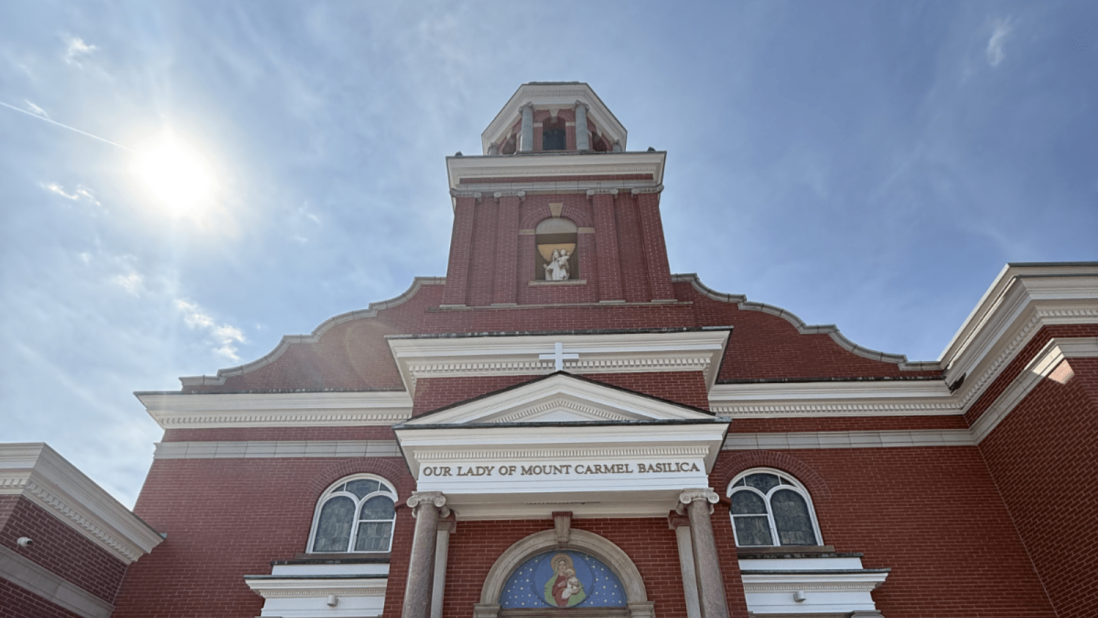 The exterior of Our Lady of Mount Carmel in Cleveland, Ohio. It is a solid brick building with a steeple leading up to a bright blue sky.