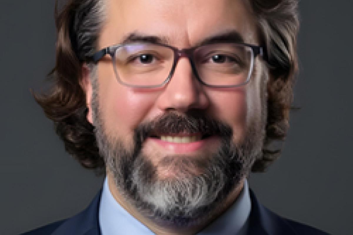 Photo of Kevin May. He has salt-and-pepper hair and a beard. He is wearing thin glasses, a navy suit jacket, a light blue shirt, and a navy tie.