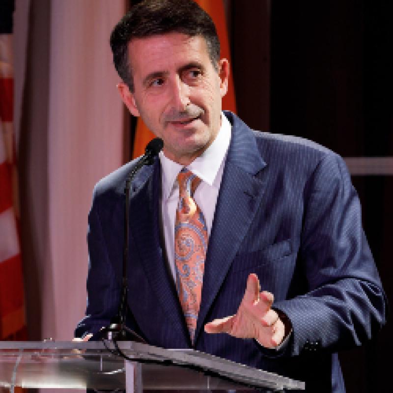 Photo of Andrew V. Abela, speaking a podium. He is wearing a navy suit and a peach tie. 