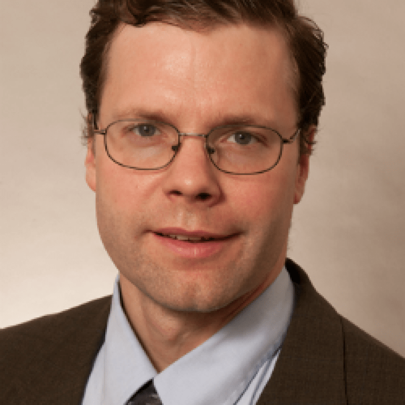 Photo of Paul Radich. He has short brown hair and thin glasses. He is wearing a brown suit with a blue shirt and brown tie.