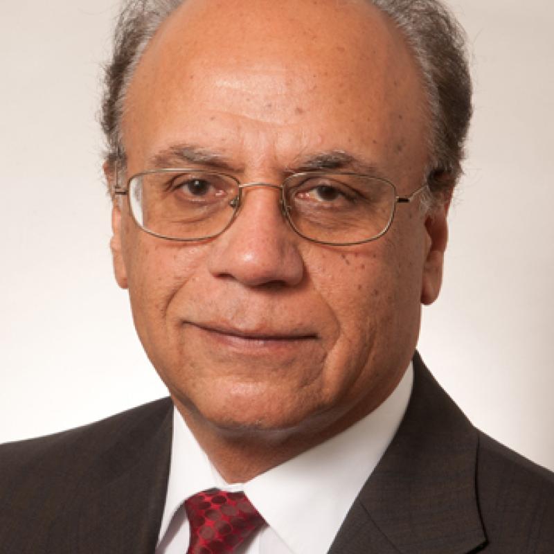 Photo of Jamshed Uppal. He has light grey hair and thin glasses. He is wearing a black suit over a white shirt and a red tie.