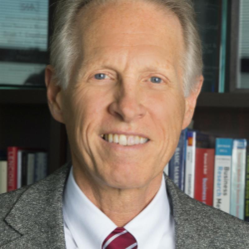 Photo of Brian Engelland. He has short white hair and is wearing a grey blazer, a white shirt, and a red and white tie.
