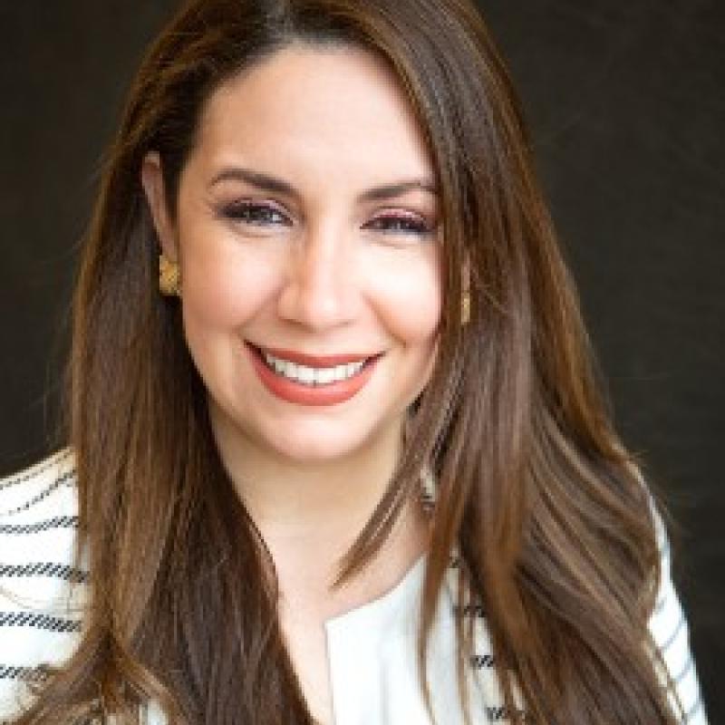 Photo of Martha Valenzuela. She has long brown hair and is wearing a white and black striped shirt.