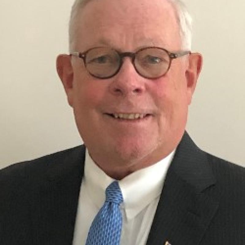 Photo of Harvey Seegers. He is wearing a black suit with a white shirt and light blue tie. He has an American flag pin on his lapel. He has white hair and round glasses.