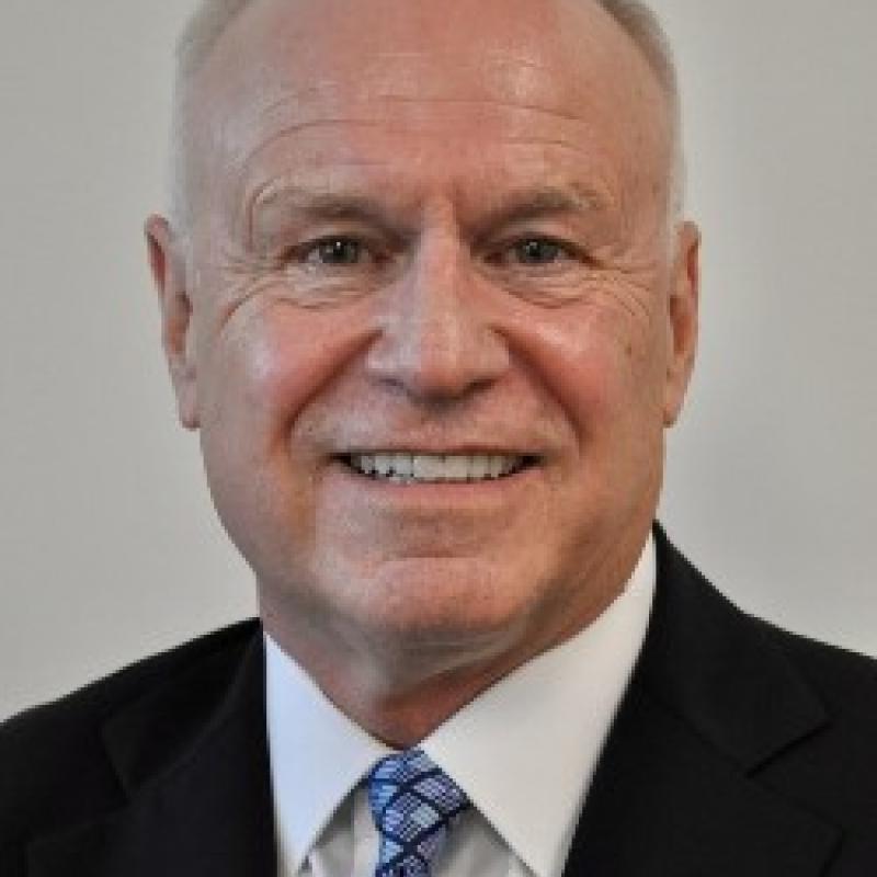 Photo of James P. Kelly. He has short white hair and is wearing a navy suit, a white shirt, and a blue tie.