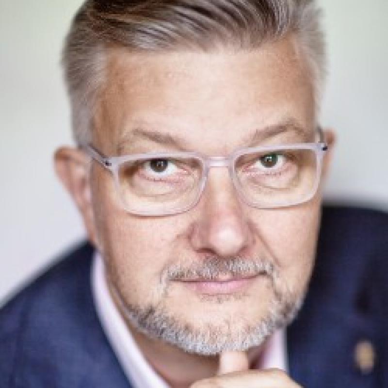 Photo of Andreas Widmer. He has short grey hair and thick glasses. He is wearing a navy suit jacket and pink shirt.