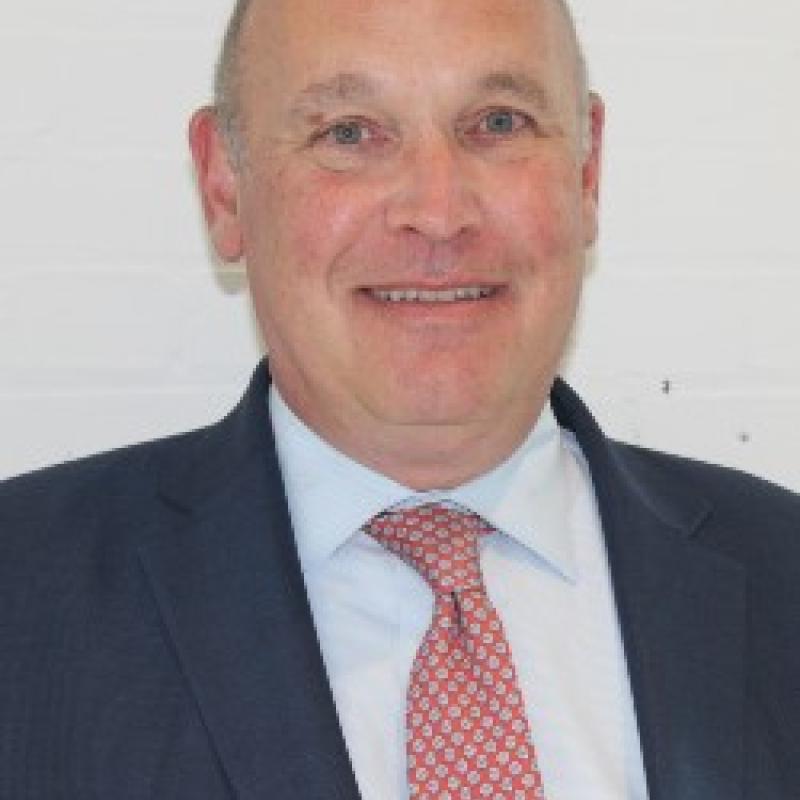 Photo of Frank Kelly. He is wearing a navy suit over a light blue shirt with a red and white tie.