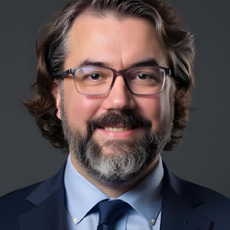 Photo of Kevin May. He has salt-and-pepper hair and a beard. He is wearing thin glasses, a navy suit jacket, a light blue shirt, and a navy tie.