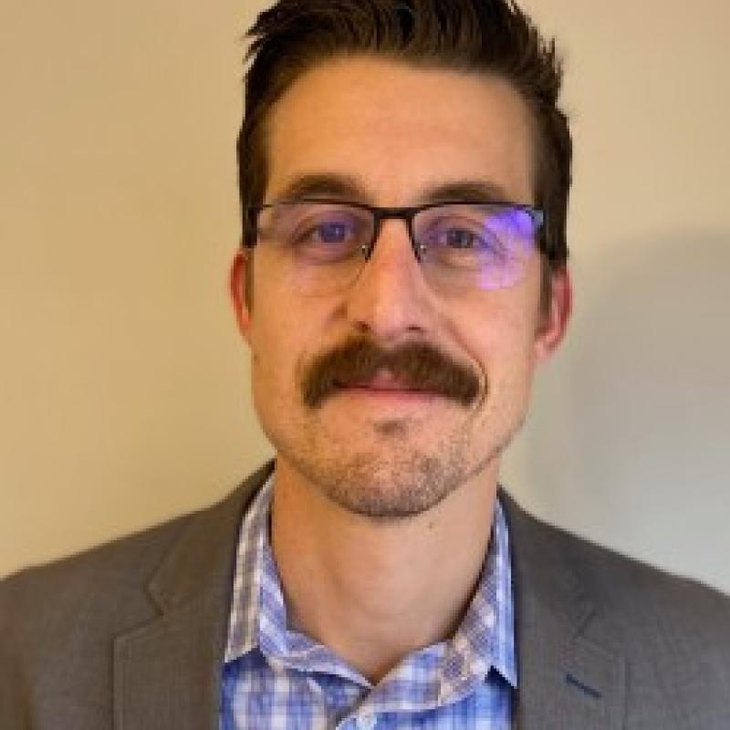 Photo of Erik W. Matson. He has dark hair and a mustache. He is wearing a grey suit jacket over a blue and white checkered shirt.