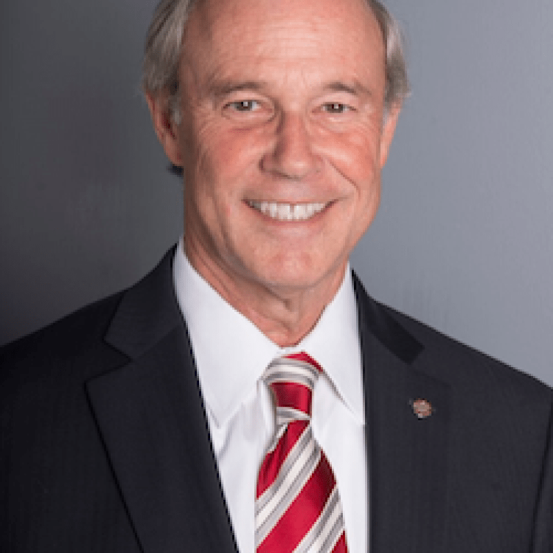 Photo of Stewart McHie. He is wearing a black suit, a white shirt, and a red and white tie. He has short grey hair.