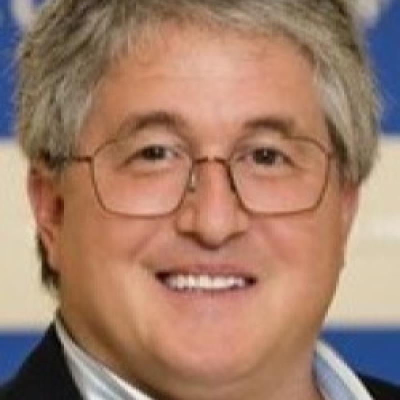 Photo of Michael Troilo. He is wearing thin glasses and has grey hair. He is wearing a navy suit jacket over a blue striped shirt.