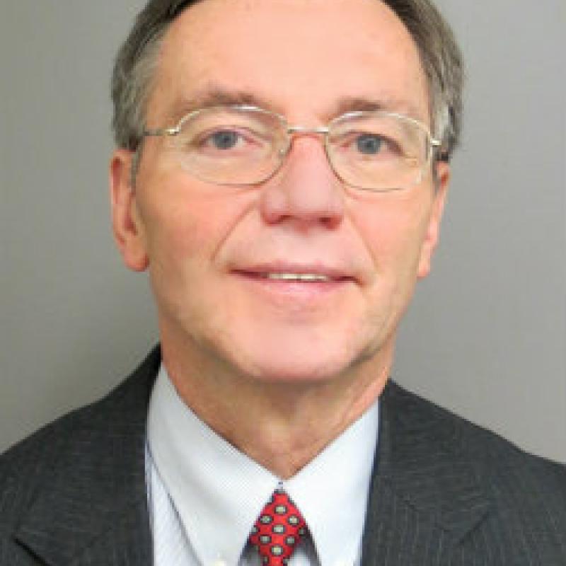 Photo of Mark Michalski. He is wearing a grey suit with a white shirt and a red spotted tie. He has short grey hair and white glasses.