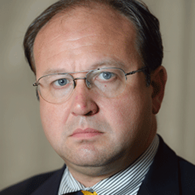 Photo of Michael New. He is wearing a navy suit, a striped shirt, and a yellow and black tie.