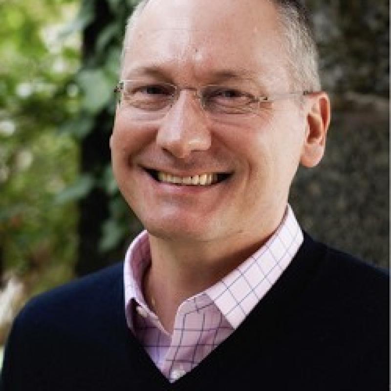 Photo of Frederic Sautet. He has salt and pepper hair and thin glasses. He is wearing a pink checked shirt under a navy sweater.