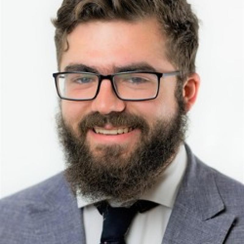 Photo of Marcus Shera. He has brown hair and a beard. He is wearing square glasses, a grey suit, and a white shirt with a black tie.