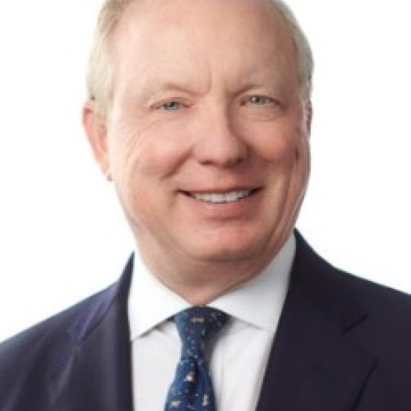 Photo of John Mino. He is wearing a navy suit, a white shirt, and a blue patterned tie. He has short grey hair.