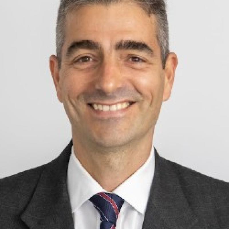 Photo of Lucas E. Well. He has short salt and pepper hair. He is wearing a black suit with a white shirt and a blue and red tie.