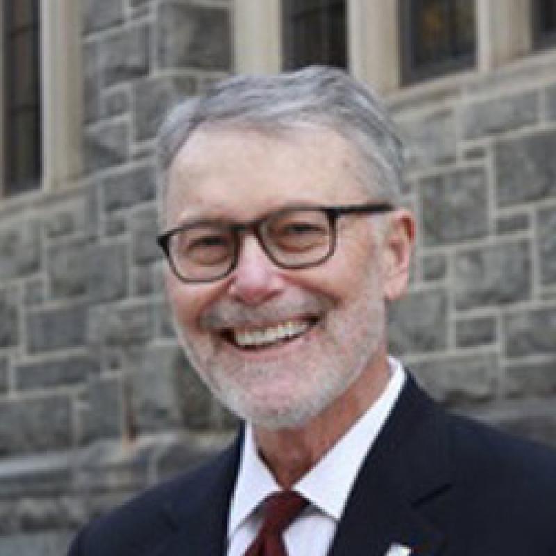 Photo of John Wesley Yoest, Jr. He has short grey hair. He is wearing a navy suit jacket, white shirt, and red tie. He has thick black glasses.