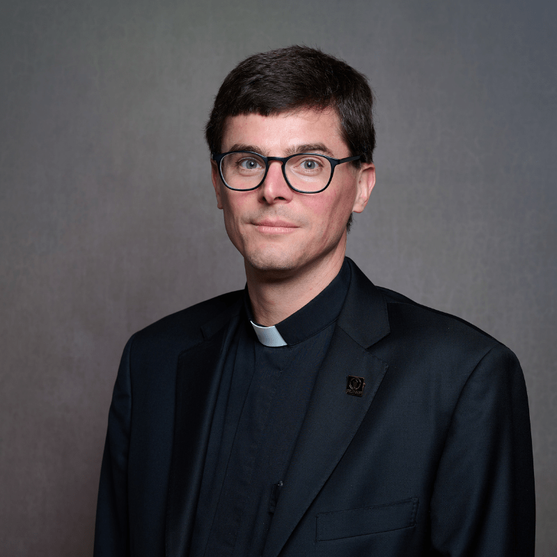 Photo of Rev. Anthony J. Stoeppel. He has brown hair and is wearing round glasses. He is wearing a black suit jacket and a black shirt with a white clerical collar.