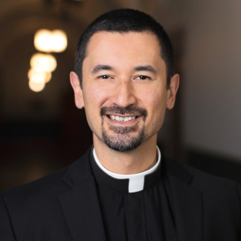 Photo of Fr. Ryan Ruiz . He is wearing a black suit jacket, a black shirt, and a clerical collar. He has dark hair and a beard.