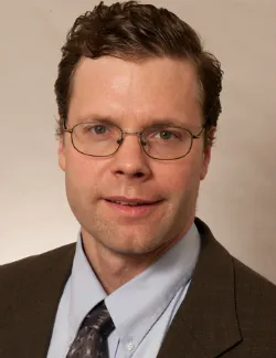 Photo of Paul Radich. He has short brown hair and thin glasses. He is wearing a brown suit with a blue shirt and brown tie.