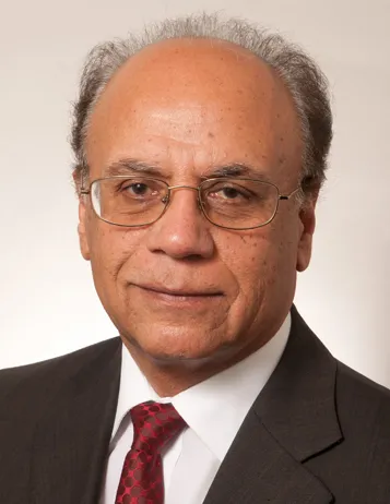 Photo of Jamshed Uppal. He has light grey hair and thin glasses. He is wearing a black suit over a white shirt and a red tie.