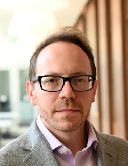 Photo of Anthony P. Cannizzaro. He has short brown hair, thick black glasses, and is wearing a grey shirt over a pink shirt.