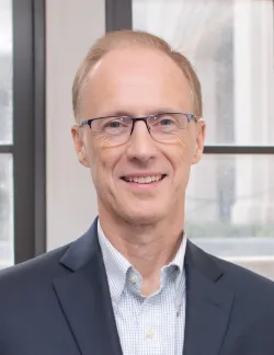 Photo of John Egan. He has short light brown hair, thin glasses, and is wearing a light blue shirt under a navy blazer.