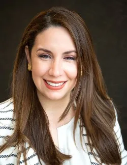 Photo of Martha Valenzuela. She has long brown hair and is wearing a white and black striped shirt.