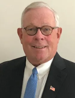 Photo of Harvey Seegers. He is wearing a black suit with a white shirt and light blue tie. He has an American flag pin on his lapel. He has white hair and round glasses.