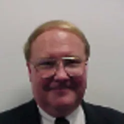 Photo of Leon Hutton. He has short blonde hair, thin glasses, and is wearing a black suit. 