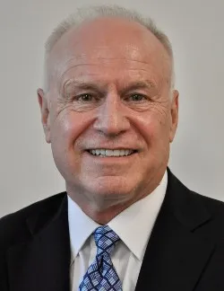 Photo of James P. Kelly. He has short white hair and is wearing a navy suit, a white shirt, and a blue tie.