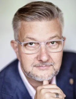 Photo of Andreas Widmer. He has short grey hair and thick glasses. He is wearing a navy suit jacket and pink shirt.