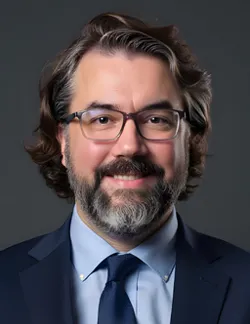 Photo of Kevin May. He has salt-and-pepper hair and a beard. He is wearing thin glasses, a navy suit jacket, a light blue shirt, and a navy tie.