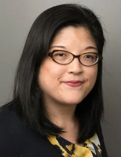 Photo of Irene Kim. She has should-length black hair. She has cat-eye glasses and is wearing a black and yellow blouse.