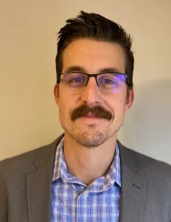 Photo of Erik W. Matson. He has dark hair and a mustache. He is wearing a grey suit jacket over a blue and white checkered shirt.