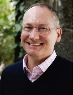 Photo of Frederic Sautet. He has salt and pepper hair and thin glasses. He is wearing a pink checked shirt under a navy sweater.