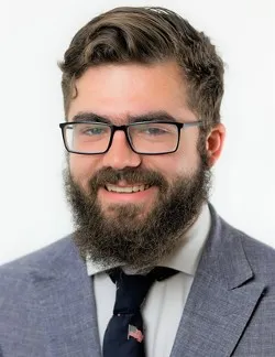 Photo of Marcus Shera. He has brown hair and a beard. He is wearing square glasses, a grey suit, and a white shirt with a black tie.