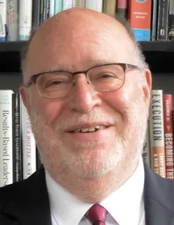 Photo of Max Torres. He wears thin glasses as well as a black suit, a white shirt, and a red tie.