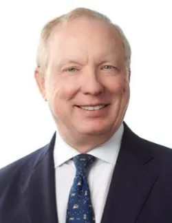 Photo of John Mino. He is wearing a navy suit, a white shirt, and a blue patterned tie. He has short grey hair.