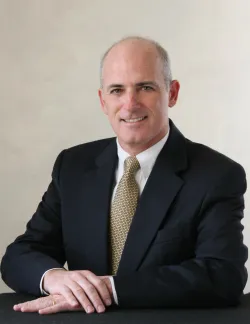 Photo of Mark Weber. He is wearing a black suit, a white shirt, and a gold tie.