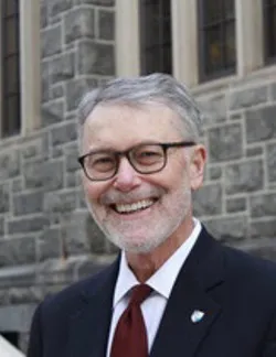 Photo of John Wesley Yoest, Jr. He has short grey hair. He is wearing a navy suit jacket, white shirt, and red tie. He has thick black glasses.