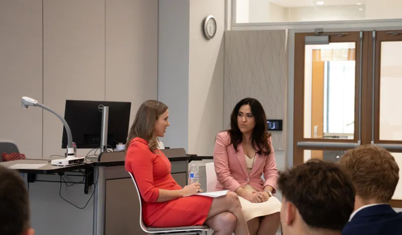 Two women are speaking to each other during a conversation in a classroom. One is wearing a red dress, another is wearing a pink blazer over a white dress.