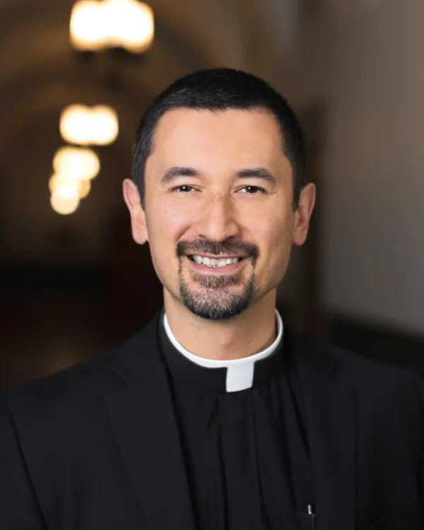 Photo of Fr. Ryan Ruiz . He is wearing a black suit jacket, a black shirt, and a clerical collar. He has dark hair and a beard.
