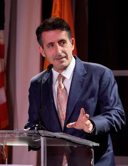 Photo of Andrew V. Abela, speaking a podium. He is wearing a navy suit and a peach tie. 