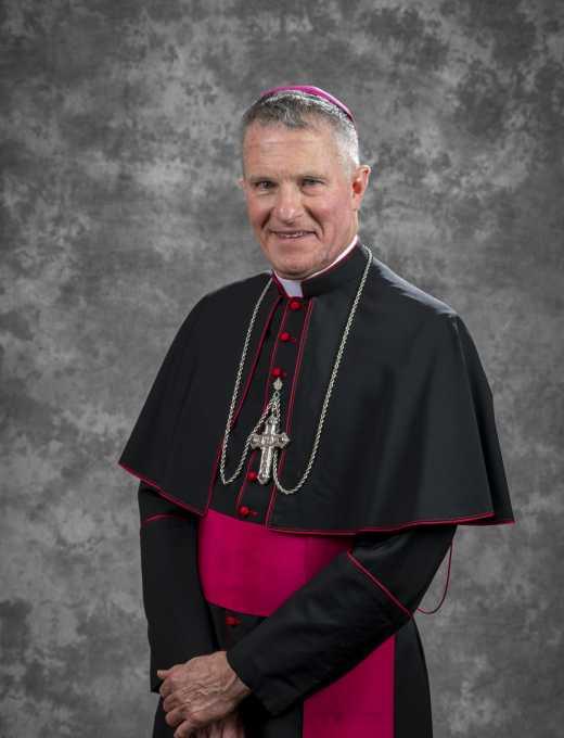 Photo of Archbishop Broglio. He is against a grey background and is wearing a black priestly garb with red accents.